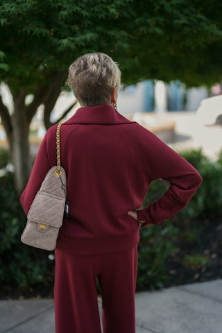 Back view from the waist up showcasing the wide folded collar, smooth knit finish, and structured raglan seams of the burgundy pullover top. Displays the shoulder fit and rear placement of the taupe quilted suede shoulder bag with a woven gold chain strap.
