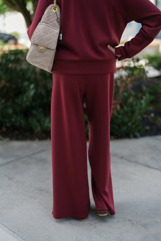 Back view from the neck down showcasing the relaxed fit and clean lines of the burgundy pullover sweatshirt and wide-leg pants. Highlights the smooth rear waistband, wide drape of the trouser legs over brown suede shoes, and the rear view of the taupe quilted crossbody bag.