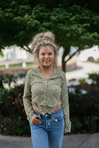 Mid-length front view highlighting the scalloped V-neckline and pearl button-down placket of the sage green floral lace top. Paired with light-to-medium wash Good American straight-leg denim jeans with one hand tucked into the pocket.