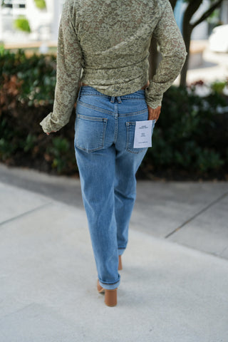 Back view showcasing the fit of the sage green sheer lace top and blue Good American Vintage Straight Ankle jeans. Highlights the back pocket design with brand tag detailing, straight-leg cut, rolled cuffs, and tan block-heel boots.