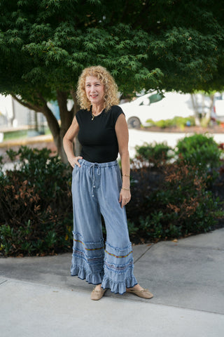 Woman posing outdoors in light wash wide-leg denim chambray pants featuring a high elastic drawstring waist and tiered ruffle hems adorned with colorful embroidered trim and raw-edge fraying. Styled with a sleeveless black top, gold jewelry, and beige ballet flats in front of lush green foliage.