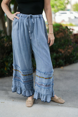 A detailed close-up shot highlighting the elastic waist with an adjustable drawstring tie and the bohemian multi-tiered hem design on the light wash wide-leg pants. Features horizontal rows of frayed denim trim, vibrant geometric embroidery, and a ruffled bottom hem paired with neutral woven flats.