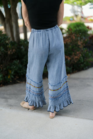 Back view from the waist down showcasing the smocked elastic waistband and the relaxed fit of the light blue wide-leg chambray trousers. Displays the continuous embroidered bands, raw-edge frayed trim, and ruffled hem detail along the back of each leg.