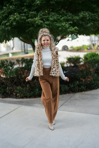 Woman posing outdoors in a plush faux fur leopard print vest styled open over an off-white ribbed turtleneck sweater and paired with high-waisted camel brown suede wide-leg pants and pointed-toe cream boots. Styled with a high curly ponytail and gold hoop earrings in front of a lush green tree backdrop.