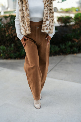 A detailed front shot from the bust down highlighting the rich texture of the high-rise camel suede barrel-leg trousers with contrast topstitching, zip fly, and metallic button closure. Shows the open drape of the soft spotted faux fur vest and off-white ribbed knit top over pointed cream booties.