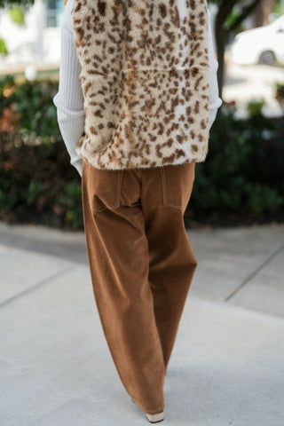 Back view from the neck down showcasing the plush plush texture and spotted pattern across the back of the leopard faux fur vest. Highlights the relaxed wide-leg fit, rear patch pockets with contrast stitching, and smooth drape of the camel brown suede pants.