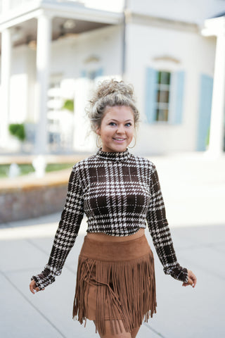 Portrait view highlighting the detailed velvet texture and plaid houndstooth-style pattern of the brown mock neck long-sleeve crop top. Paired with a high-waisted chestnut faux suede skirt featuring a wide waistband and tiered fringe trim.