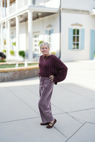 Woman posing outdoors in vintage-washed plum purple corduroy wide-leg trousers, styled with a deep burgundy open-knit crochet crewneck sweater and brown suede Mary Jane flats. The high-waisted pants feature statement front patch pockets with delicate scalloped trim details. Styled with a chunky gold chain necklace in front of a white building with light blue shutters.