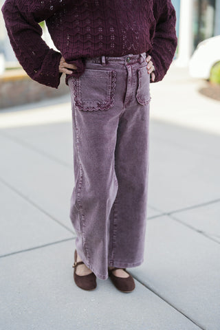 A detailed close-up shot highlighting the fine-ridge corduroy texture and decorative scalloped edges on the front patch pockets of the dusty mauve wide-leg pants. Shows the zip fly, metallic top button, structured waistband, and relaxed wide-leg drape over dark brown buckled suede flats.