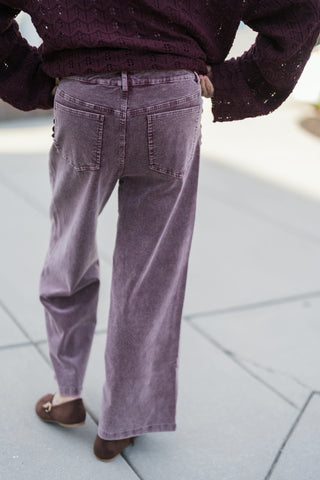 Back view from the waist down demonstrating the fit and silhouette of the washed purple corduroy pants. Features traditional back patch pockets, vertical ribbed corduroy texture, and a wide-leg hem worn with brown suede Mary Jane shoes beneath a cropped burgundy sweater.