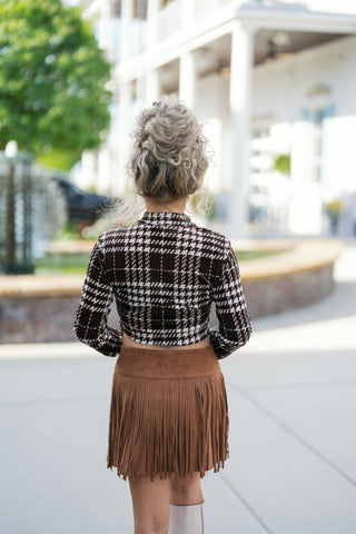 Back view showcasing the back coverage of the brown and white plaid cropped long-sleeve top and the straight hemline with hanging fringe on the faux suede mini skirt. Features the back profile of the model's curly hair updo and tall cream boots.