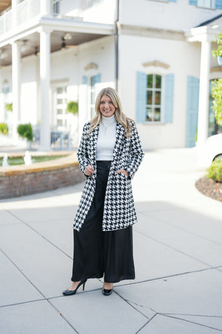 Woman posing outdoors in a black and white houndstooth open-front mid-length overcoat, styled with a fitted white ribbed turtleneck sweater, high-waisted black wide-leg trousers, and black patent heels. The tailored coat features notch lapels, deep side pockets, and a mid-thigh hemline. Styled in front of a white building with light blue shutters.