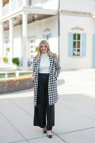 Full-length front view showcasing the black and white houndstooth coat layered open over a tucked-in white turtleneck and black high-waisted wide-leg trousers with front waist buttons. The model stands on a concrete plaza with her hands resting in the coat pockets.