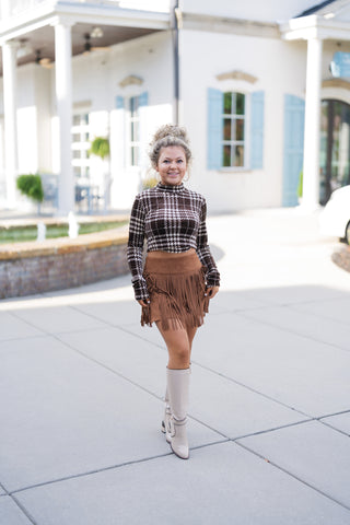 Woman posing outdoors in a brown and cream plaid mock neck long-sleeve crop top paired with a cognac brown faux suede fringe mini skirt and off-white knee-high boots. Styled with gold hoop earrings and her curly hair in an updo in front of a white building background.