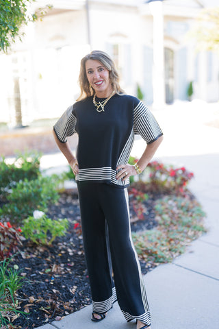 A woman posing outdoors on a sunny sidewalk with her hands on her hips, wearing a chic two-piece knit set featuring a short-sleeve black top and wide-leg trousers. The relaxed-fit crewneck sweater top features contrasting cream-and-black vertical striped panels down the side seams and sleeves, finished with horizontal striped border trim along the hem. The look is completed with matching side-striped wide-leg knit pants, black open-toe heel sandals, and a gold chunky chain necklace.