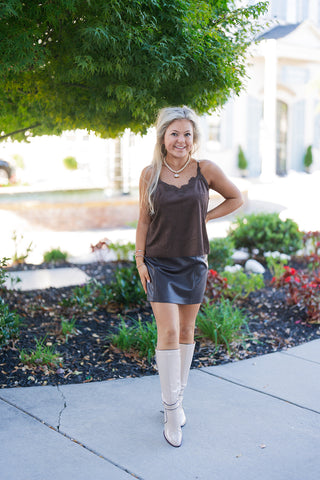 A woman posing outdoors on a sidewalk wearing a dark chocolate brown faux suede camisole top paired with a matching dark brown faux leather mini skirt. The cami features delicate lace trim along the V-neckline and thin adjustable spaghetti straps. Styled for a fall fashion look with cream patent knee-high boots, a short beaded pendant necklace, gold hoop earrings, and stacked gold bracelets.