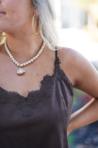 Close-up cropped view focusing on the intricate lace trim along the neckline of the rich brown suede-textured camisole. Demonstrates the delicate eyelash lace edge along the V-neck cut and subtle bust darts, paired with a beaded necklace featuring a gold-rimmed pendant.