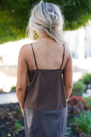 Back view of the dark brown faux suede camisole showing the straight square back hem, fine adjustable spaghetti straps, and clean hemline. Paired with the back of a dark faux leather mini skirt featuring visible vertical seam stitching, set against a blurred green park backdrop.