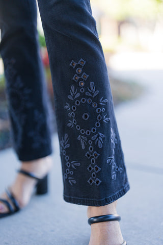 Detailed close-up of the lower leg showing the black cropped flare denim jean with intricate tonal thread embroidery, geometric patterns, and subtle stud embellishments framing the hem above black strappy heels.