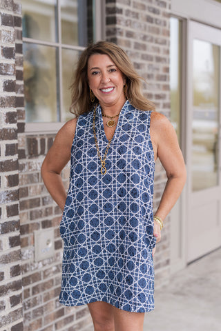 A medium shot of the Nantucket Lattice Dress highlighting the intricate, textured woven fabric and the "lattice" geometric design. The V-neckline is accented with a gold coin pendant and a toggle link necklace. The model is smiling with her hands in her pockets, showcasing the dress's practical and polished coastal aesthetic.