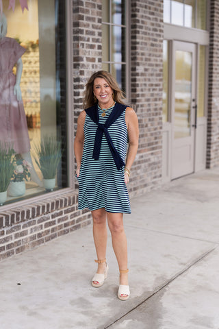 A full-length view of a woman posing in the Tied & True Stripe Dress, a preppy navy and mint green horizontal striped sleeveless mini dress. The dress features a unique "built-in" navy knit scarf detail draped over the shoulders and tied at the chest for a layered look. Styled with cream platform wedge sandals, gold bangles, and a coin necklace in front of a modern brick building.