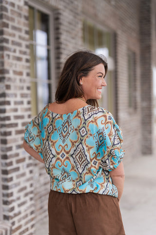 Rear and side view of the Ikat Inkscape Blouse highlighting the effortless drape of the lightweight fabric. This shot showcases the short dolman sleeves cinched with elastic cuffs to create a subtle balloon effect. The bold, multi-tonal ikat pattern flows seamlessly across the back, emphasizing the breezy, oversized fit against a neutral brick storefront.