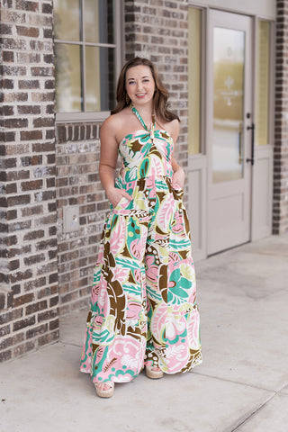 Woman posing in the Ibiza Icon Jumpsuit, a vibrant multi-colored piece featuring a bold tropical floral print in lime green, bubblegum pink, and earthy bronze. The design showcases a chic twist-front halter neckline and dramatic wide tiered legs that create a voluminous, dress-like silhouette. She is styled with beige platform sandals in front of a modern brick boutique, highlighting the jumpsuit's functional side pockets.