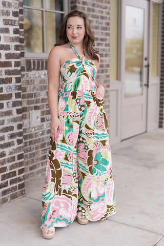 A portrait view of the Ibiza Icon Jumpsuit focusing on the sophisticated twist-front halter neckline and the intricate botanical pattern. The vibrant pink and green floral tones pop against the crisp white background. The model’s relaxed pose emphasizes the secure fit of the bodice and the premium, breezy feel of the seasonal fabric
