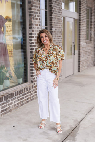 A woman posing in the Amber Garden Blouse, a toffee-colored top featuring an oversized paisley and floral motif in sage, vanilla, and sky blue. She has styled the blouse tucked into high-waisted white wide-leg trousers with raw hems and beige block-heeled sandals. The shot is taken outdoors on a sidewalk in front of a brick boutique with a mannequin visible in the window.