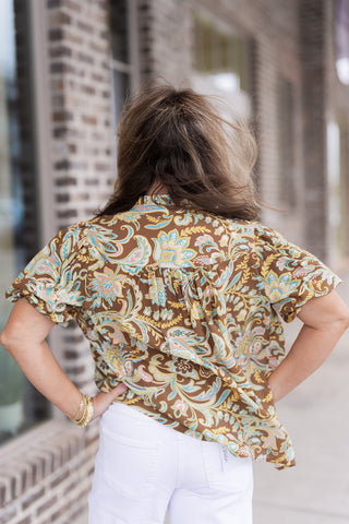 Rear view of the Amber Garden Blouse showcasing the consistent flow of the multi-colored floral and paisley print across the shoulders and back. This angle emphasizes the relaxed, flowy fit of the bodice and the soft gathering at the yoke. The model stands with her hands on her hips, highlighting the volume of the elasticated puff sleeves.
