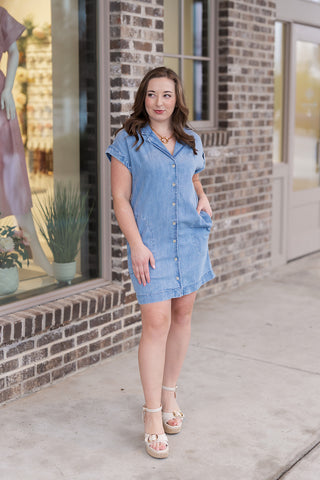Woman posing in the Z Supply Napa Mini Dress, a light-wash denim shirtdress crafted from a premium 63% Cotton and 37% Lyocell blend. The design features a classic pointed collar, a functional button-front closure, and discreet side seam pockets. This relaxed mini-length silhouette is treated with a heavy soft wash for an ultra-soft feel and is styled with ivory platform sandals.