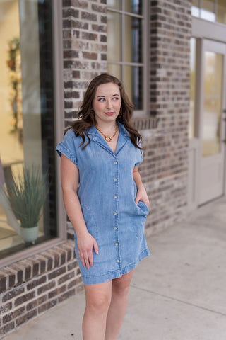 Woman posing in the Z Supply Napa Mini Dress, a light-wash denim shirtdress crafted from a premium 63% Cotton and 37% Lyocell blend. The design features a classic pointed collar, a functional button-front closure, and discreet side seam pockets. This relaxed mini-length silhouette is treated with a heavy soft wash for an ultra-soft feel and is styled with ivory platform sandals.