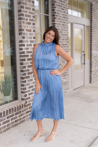 Woman posing in the Seraphina Sky Midi Dress; a romantic, dusty sky blue sleeveless midi dress featuring an intricate vertical pleated texture. The design includes an elegant high-neckline, a flattering cinched waist, and a flowing A-line skirt that creates a graceful, elongating silhouette. Styled with tan-toned heeled sandals and gold jewelry for a sophisticated garden party or evening look.