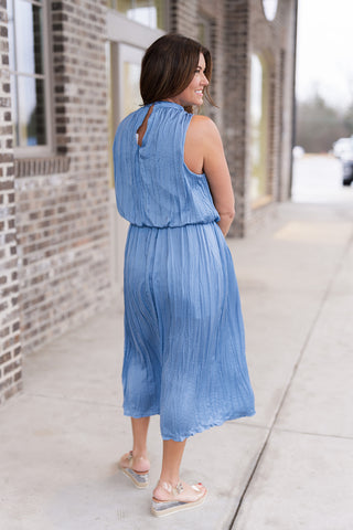 Woman posing in the Seraphina Sky Midi Dress; a romantic, dusty sky blue sleeveless midi dress featuring an intricate vertical pleated texture. The design includes an elegant high-neckline, a flattering cinched waist, and a flowing A-line skirt that creates a graceful, elongating silhouette. Styled with tan-toned heeled sandals and gold jewelry for a sophisticated garden party or evening look.