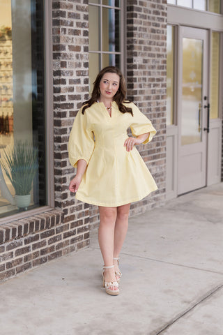 A woman posing in the Lemon Drop Mini Dress, a vibrant lemon yellow dress featuring a structured A-line silhouette. The design includes architectural elbow-length puff sleeves with pleated cuff details and a modern split V-neckline. She is styled with cream platform espandrill sandals and gold jewelry in front of a brick storefront.