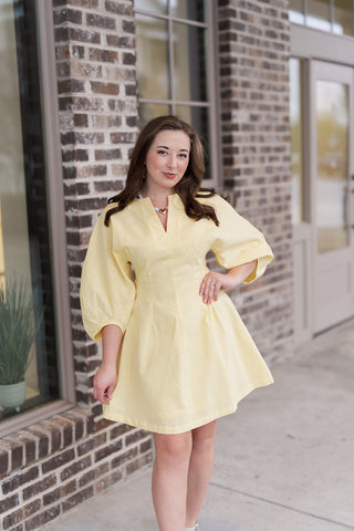 A portrait view of the Lemon Drop Mini Dress highlighting the high-quality textured woven fabric and the tailored waist darting that creates a figure-flattering fit. The split V-neckline is accessorized with a gold heart pendant necklace, emphasizing the dress's clean bodice and playful, boutique-inspired aesthetic.