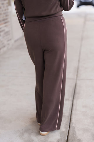 Rear view of the Fast Track Joggers showcasing the clean, effortless drape of the wide-leg silhouette. The chocolate brown fabric is accented by continuous light pink piping detail, offering a streamlined aesthetic that transitions seamlessly from studio to street.