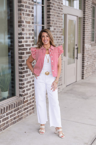 Woman posing in the Regatta Ruffle Vest, a red and white vertical striped button-down vest featuring dramatic, multi-layered ruffled cap sleeves. She styles it over a white bodysuit with white high-waisted wide-leg jeans, a gold statement buckle belt, and cream platform sandals for a high-fashion nautical aesthetic.