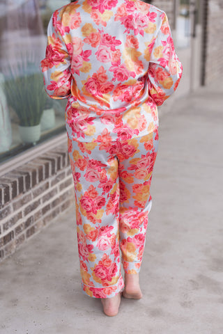 Back view of a woman wearing the Sweet Briar Pajama Set, emphasizing the all-over floral rose pattern and the clean, tailored drape of the long-sleeve button-up and matching pants. The lightweight fabric features a notched collar with delicate piping, set against a blurred storefront background.
