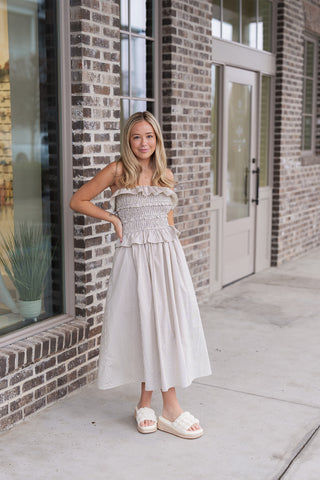 Woman wearing the Sunlit Stripe Midi Dress, a strapless strapless silhouette featuring a classic vertical striped pattern. The dress highlights a fully smocked bodice for a customized fit with romantic, layered ruffle trims at the neckline and waist. The flowy A-line midi skirt is crafted from a breathable cotton-blend material, styled with cream-colored woven platform slides for a coastal summer look.