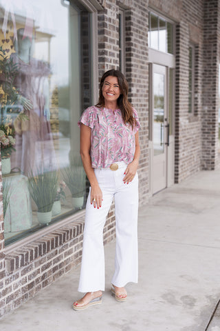 Woman posing in the Amethyst Air Blouse, a lavender-toned top featuring an oversized leaf print in amethyst, sage, and cream. She has styled the blouse with high-waisted white wide-leg trousers, a white belt with a large gold buckle, and clear platform wedge sandals in front of a modern brick boutique window.