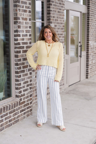 A woman posing in the Lemon Sherbert Top, a butter-yellow long-sleeve sweater featuring an intricate open-knit crochet pattern. She is paired with high-waisted gray and white vertical striped trousers and cream platform sandals, creating a bright and airy spring aesthetic.