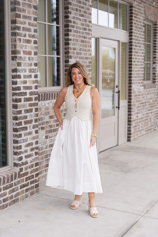 Woman posing in the Linen Louvre Midi Dress, a sophisticated mixed-media piece featuring a natural-toned linen-blend vest bodice with functional wood-toned buttons and a pointed waistcoat hem. The tailored top is seamlessly attached to a crisp white voluminous midi skirt, creating a chic two-piece illusion. Styled for a "quiet luxury" aesthetic with tan platform wedge sandals and gold jewelry in front of a modern brick storefront.