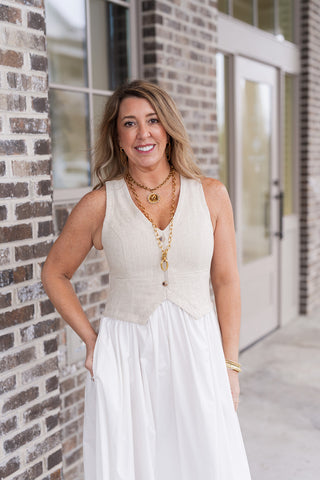 A portrait view of the Linen Louvre Midi Dress highlighting the architectural detail of the structured V-neckline vest bodice. The natural linen-blend fabric offers a rich, organic texture, complemented by layered gold coin and link necklaces. The design provides a refined, tailored fit that cinches the waist naturally while maintaining breathable, elevated style.