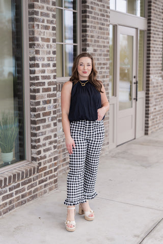 Woman posing in the Midnight Muse Tank, a sophisticated obsidian black sleeveless blouse with a high, gathered neckline. She is styled with high-waisted black and white gingham trousers featuring a playful ruffled hem and neutral platform sandals. The shot is taken in front of a modern brick boutique, showcasing the tank's elegant, slightly tapered silhouette.