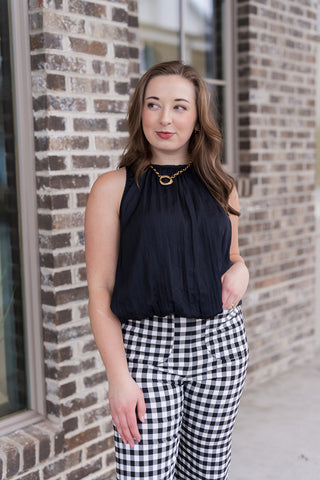 A portrait view of the Midnight Muse Tank highlighting the intricate gathering at the crew neckline, which creates a soft, fluid drape across the bodice. The deep black fabric features a subtle, light-catching texture that pairs perfectly with the featured gold oval-link necklace. This halter-inspired design provides a polished, modern look for any occasion.