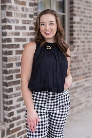 A medium shot focusing on the Midnight Muse Tank's premium fabric and clean-finished armholes. The model is looking off-camera, emphasizing the subtle blouson effect at the waist where the tank is tucked into gingham print trousers. The rich, saturated black hue of the top contrasts beautifully with her gold jewelry and the textured brick background.