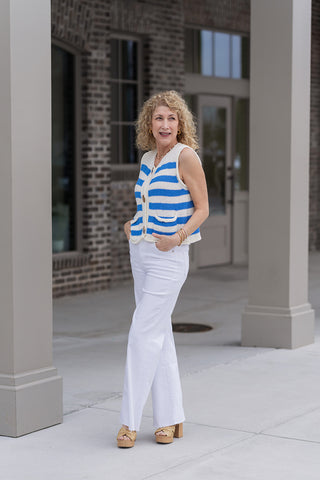 A woman modeling the Saint-Tropez Stripe Vest, a sleeveless knit piece featuring elegant cream and cornflower blue horizontal stripes. The vest is styled for a "French Riviera" aesthetic with high-waisted white wide-leg trousers and tan woven platform heels. She stands under a modern storefront awning, showcasing the vest's relaxed, slightly cropped fit.