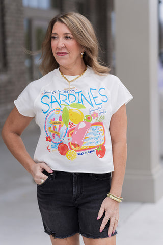 A detailed view of the Amalfi Appetizer Tee highlighting the hand-painted style graphic of colorful sardine tins and zesty lemons. The image showcases the trendy raw-edge detailing on the sleeves and hem, the raglan stitching at the shoulders, and a gold link necklace with a cross pendant.
