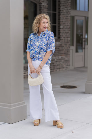 A woman stands in front of a boutique wearing the Mediterranean Muse Blouse, an exquisite short-sleeve button-down featuring an intricate navy blue paisley and botanical print on a crisp ivory backdrop. She has styled the blouse with high-waisted white trousers, tan woven platform heels, and a small cream-colored handbag for a sophisticated "Coastal Grandmother" look.